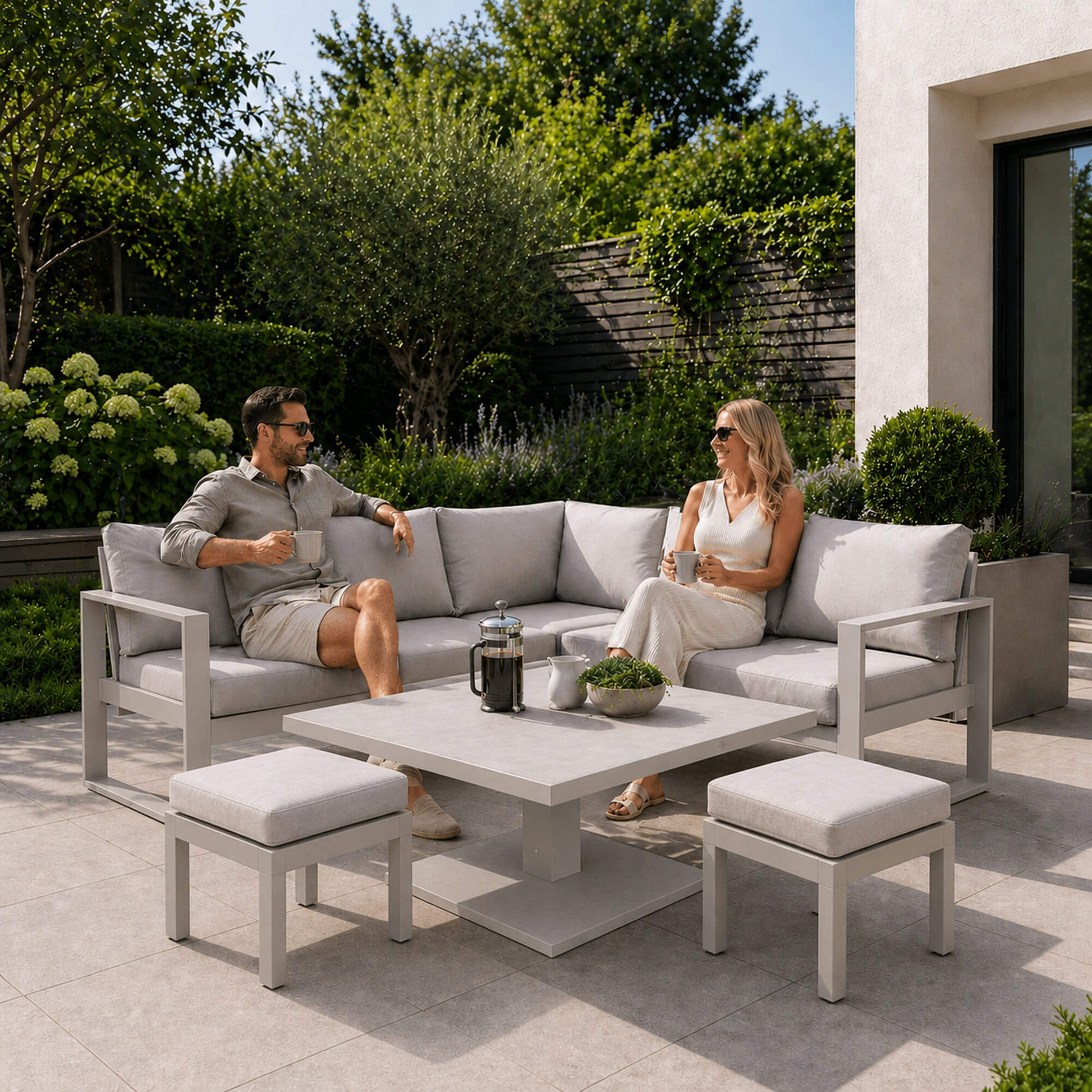 Two people sitting on a patio set with the Como stone corner set, foot stools surrounded by greenery.